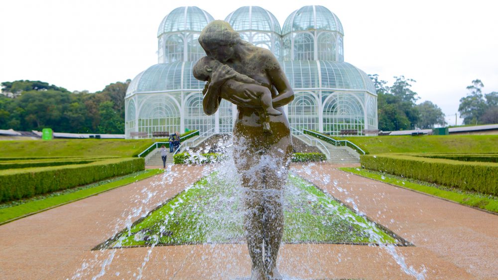 Estátua com fonte de água à frente da imagem com a estrutura de metal que abriga plantas ao fundo