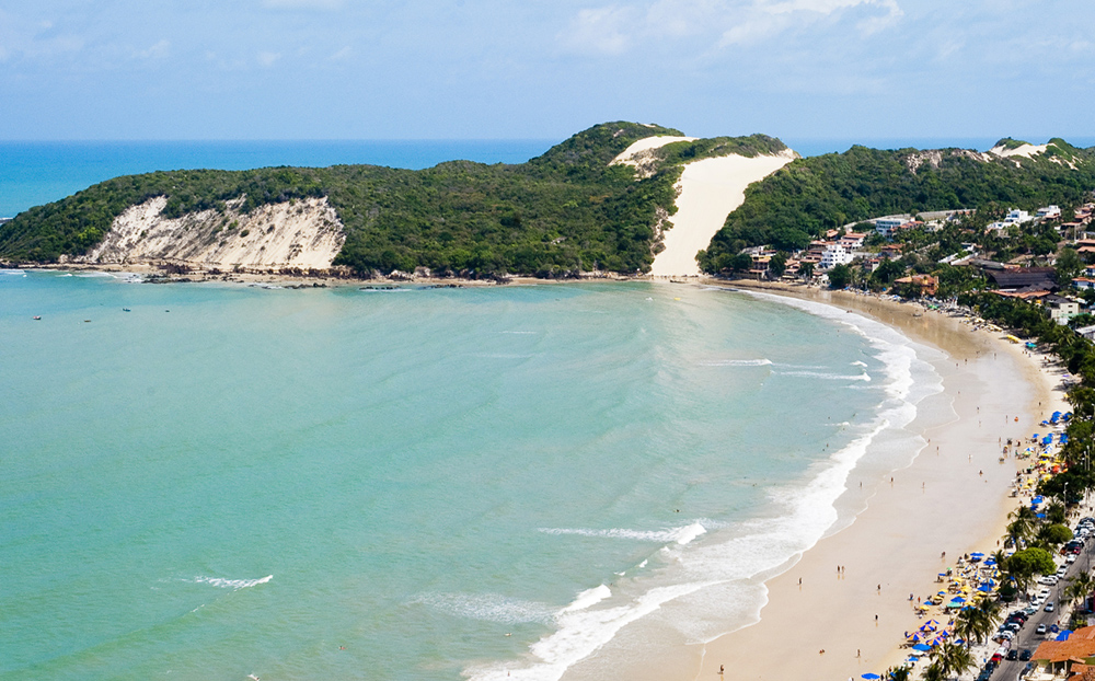 Faixa de areia e de oceano com dunas e vegetação ao fundo