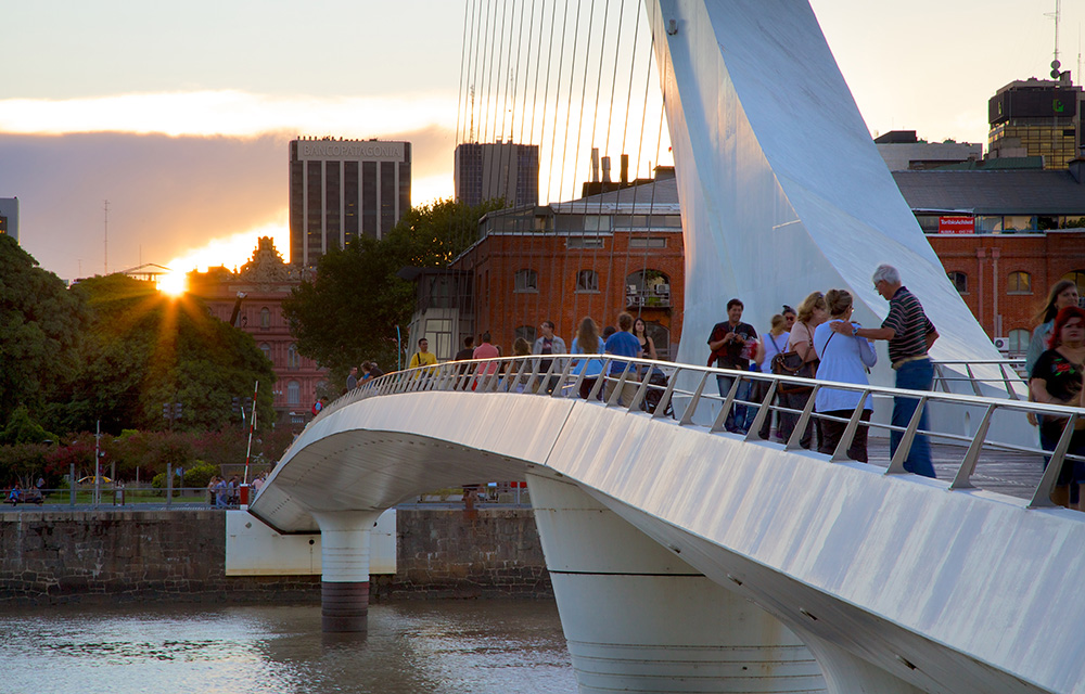 Vista lateral da Puente de la Mujer com pessoas sobre a ponte e pôr do sol ao fundo.